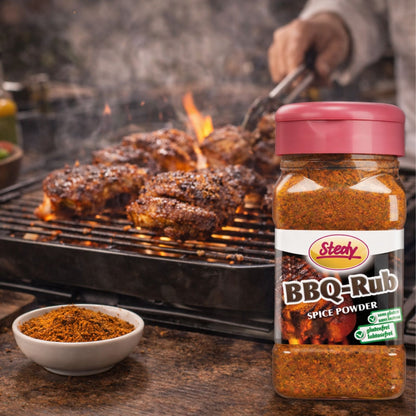 BBQ-Rub Gewürzstreuer von Stedy neben gegrilltem Fleisch auf einem Holztisch mit Rauch und Grillzange im Hintergrund